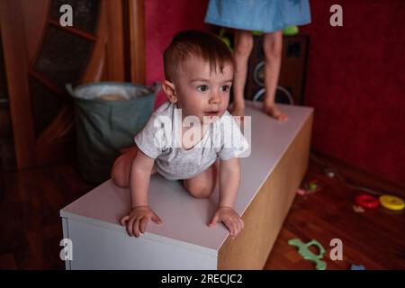 Porträt eines Kleinkindes im grauen Bodysuit, das auf einer weißen Kommode in einem Spielzeugchaos im Kinderzimmer liegt. Erziehung in Kreativität, Handlungsfreiheit. Ba Stockfoto