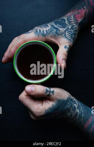 Tätowierte Hände eines Mannes, der eine Tasse Kaffee auf dunklem Hintergrund hält. Draufsicht. Vertikal Stockfoto