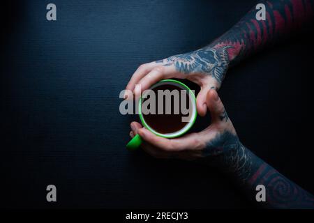 Tätowierte Hände eines Mannes, der eine Tasse Kaffee auf dunklem Hintergrund hält Stockfoto