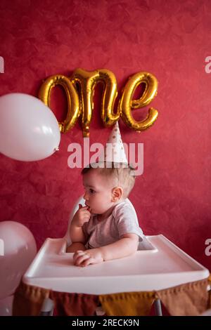 Portrait eines einjährigen Jungen mit festlichem Hut sitzt auf einem Kinderstuhl mit Stofffahnen. Kind auf isoliertem roten Hintergrund mit goldenen Luftballons aus Folie Stockfoto