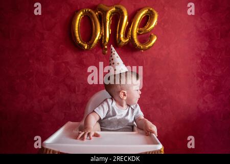 Portrait eines einjährigen Jungen mit festlichem Hut sitzt auf einem Kinderstuhl mit Stofffahnen. Kind auf isoliertem roten Hintergrund mit goldenen Luftballons aus Folie Stockfoto