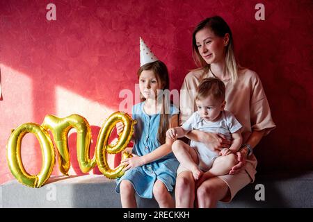 Eine junge Mutter sitzt auf dem Sofa mit kleinen Kindern in der Nähe der roten Wand auf einem grauen Sofa. Familie mit einem Elternteil feiert das erste Jahr des Babys. Die ältere Schwester ist Hure Stockfoto