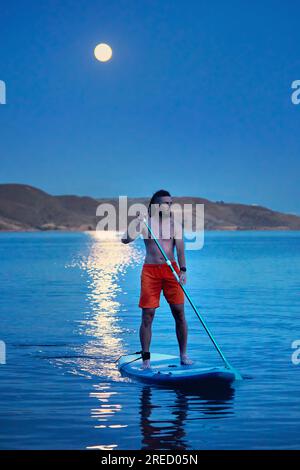 Ein Surfer in orangefarbenem Hemd auf Stand-Up-Paddle-Boards SUP in Silhouette gegen den Vollmond-Mondweg nahe der Insel am Kapchagay-See in Kasachstan. Stockfoto