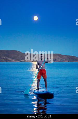 Ein Surfer in orangefarbenem Hemd auf Stand-Up-Paddle-Boards SUP in Silhouette gegen den Vollmond-Mondweg nahe der Insel am Kapchagay-See in Kasachstan. Stockfoto