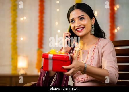 Fröhliches junges indisches Mädchen, das mit dem Handy spricht, indem es während der Festivalfeier zu Hause eine Geschenkbox in der Hand hält - Konzept von Glück, Kommunikation Stockfoto