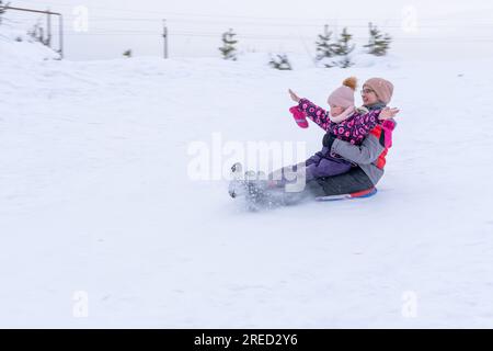 Zwei glückliche Mutter und Tochter, die auf einem Schlitten einen schneebedeckten Hügel hinunterfahren. Zwei Schwestern in Winterkleidung ruhen sich im Winter aus. Wintersport im Freien. Familie Stockfoto