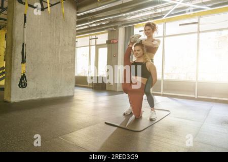 Sportliche junge Frau, die Stretching-Übungen mit Hilfe eines persönlichen Trainers im Fitnessstudio macht Stockfoto