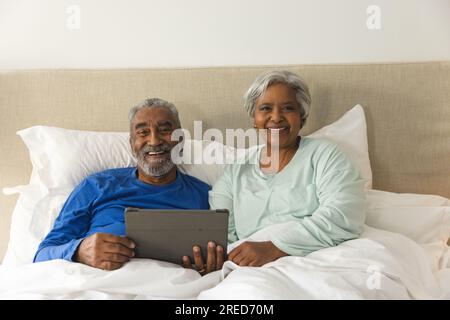 Porträt eines glücklichen älteren, birassischen Paares, das zu Hause mit einem Tablet im Bett liegt Stockfoto