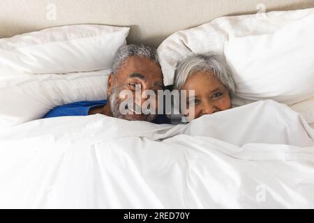 Porträt eines glücklichen älteren, birassischen Paares, das zu Hause im Bett lag und mit Bettwäsche bedeckt war Stockfoto