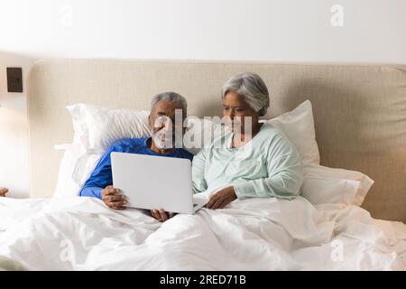 Glückliches älteres birassisches Paar, das zu Hause mit einem Laptop im Bett lag Stockfoto