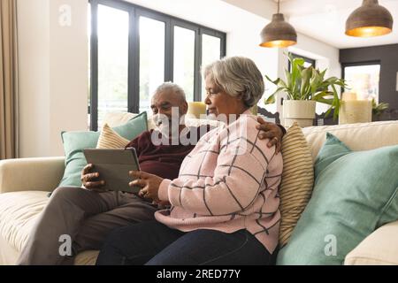 Glückliches älteres, birassisches Paar, das zu Hause auf dem Sofa sitzt und ein Tablet-Videogespräch führt Stockfoto