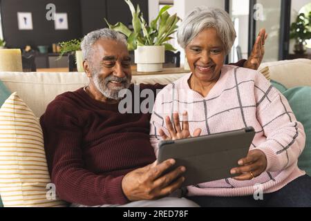 Glückliches älteres birassisches Paar, das auf dem Sofa sitzt und zu Hause ein Tablet-Videogespräch führt Stockfoto