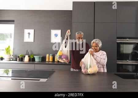 Glückliches älteres, birassisches Paar, das zu Hause Einkaufstüten in der Küche hat Stockfoto