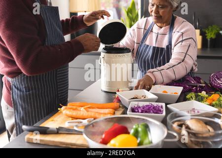 Ein älteres birassisches Paar, das Schürzen trägt und Essensabfälle in der Küche zu Hause kompostiert Stockfoto