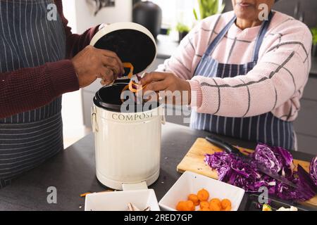 Ein älteres birassisches Paar, das Schürzen trägt und Essensabfälle in der Küche zu Hause kompostiert Stockfoto
