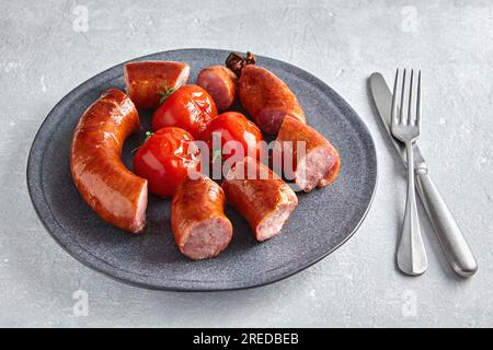 Gegrillte Wurst in Scheiben geschnitten und gebratene Tomaten auf einem grauen Keramikteller mit Besteck auf einem grauen Betontisch Stockfoto