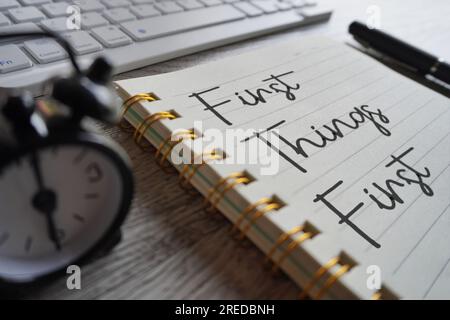 Nahaufnahme von Wecker, Tastatur und Notizbuch mit Text FIRST THINGS FIRST. Stockfoto