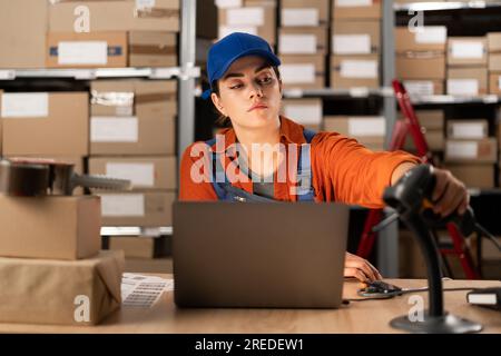 Junge Mitarbeiterin, die mit einem Laptop arbeitet und einen Strichcode-Scanner im Lagerlager verwendet. Menschen-, Lager- und Industriekonzept Stockfoto