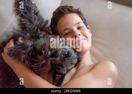 Glücklich lächelnder Junge auf dem Bett liegend und lachend in der Kamera mit seinem Hund. Konzept von glücklichen Kindern, die gute Zeit zu Hause haben. Stockfoto