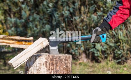 Spaltaxt in der menschlichen Hand und fliegende Holzstämme oder Sägemehl auf unscharfem grünen Naturhintergrund. Nahaufnahme der Arbeit mit Axt bei der Herstellung von Holzbrennstoff. Stockfoto