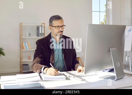 Porträt eines lächelnden, grauhaarigen Geschäftsmanns mit Brille, der vor seinem Computerbildschirm in einem modernen, hellen und geräumigen Büro sitzt. Stockfoto