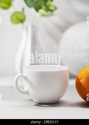 Weißer Keramikbecher und Orange auf dem Hintergrund verschwommener Küchenutensilien auf dem Tisch, Vorlage für den Designer Stockfoto