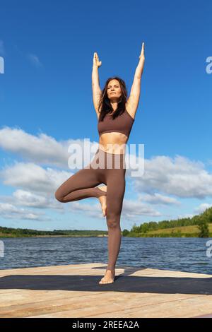 Yoga-Praktizierende Frau führt Vrikshasana-Training durch, Baum-Pose mit erhobenen Armen, Asana balancieren, sportliches Training am Ufer des Sees Stockfoto
