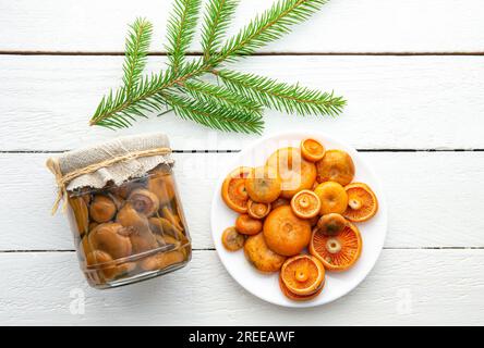 Marinierte Pilze, orangefarbene Milchkappe oder falsche Safranmilchkappe, Lactarius Deterrimus in einem Glasgefäß und frisch gepflückte Pilze auf dem Teller, weißes Holz. Stockfoto