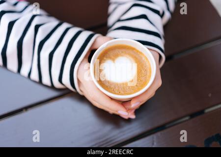 Nahaufnahme von oben, von einer unbekannten jungen Frau, die eine heiße Tasse Kaffee in Herzform hält. Nahaufnahme von unbekannten weiblichen Fingern Stockfoto