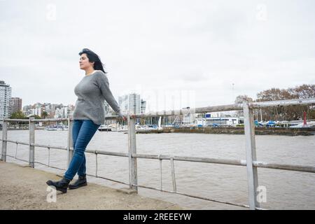 Junge abenteuerlustige Touristenlatina in einem grauen Pullover und einer blauen jeans-Hose, steht auf dem Geländer des Piers über dem Fluss und besucht Stockfoto