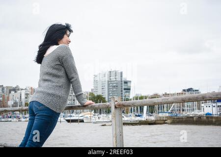 Junge abenteuerlustige Touristenlatina in einem grauen Pullover und einer blauen jeans-Hose, steht auf dem Geländer des Piers über dem Fluss und besucht Stockfoto