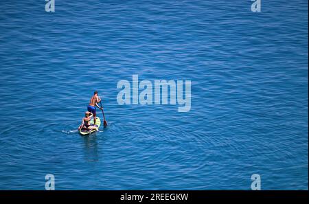 Ein junges weißes Paar paddelt auf dem ruhigen blauen Meer - ein Mann steht und schiebt ein Ruder ins Wasser, eine Frau sitzt auf dem Brett - High-an Stockfoto