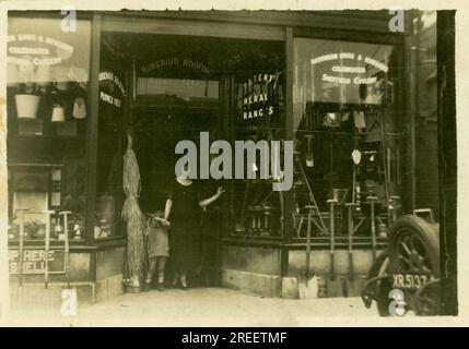 Maurice A. Clarke Ironmongers/Eisenwarenladen aus den 1920er Jahren – die Frau der Eigentümerin steht mit ihrem Kind in einer Tür. Vor dem Laden steht der Familienwagen Morris Cowley, die „Bullnose“, für 2 Personen geöffnet. Die Rückseite ist nur mit dem Buchstaben XR zu sehen, der auf eine Londoner Zulassung hinweist, Großbritannien Ungefähr 1925 Stockfoto