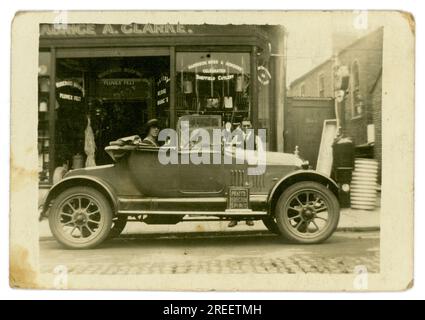 Original 1920 Jahre kleine Postkarte Maurice A. Clarke Ironmongers/Eisenwarenladen - kleines Familienunternehmen - die Frau des Eigentümers sitzt im Auto, Morris Cowley 'Bullnose', 2-Sitzer, möglicherweise zeigt sie das neue Familienauto. London, Großbritannien Ungefähr 1925. Stockfoto