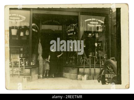 Maurice A. Clarke Ironmongers/Eisenwarenladen aus den 1920er Jahren – die Frau der Eigentümerin steht mit ihrem Kind in einer Tür. Vor dem Laden steht der Familienwagen Morris Cowley, die „Bullnose“, für 2 Personen geöffnet. Die Rückseite ist nur mit dem Buchstaben XR zu sehen, der auf eine Londoner Zulassung hinweist, Großbritannien Ungefähr 1925 Stockfoto