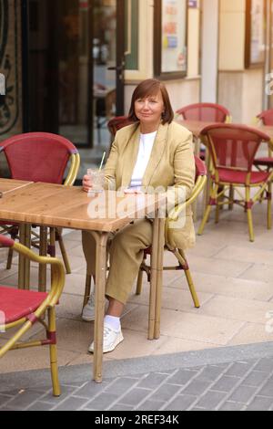 Attraktive, stilvolle, reife Frau 50-60 mit kurzen Haaren in einer gelben Jacke trinkt Kaffee, Milch, trinkt mit Stroh und entspannt im Café in Europa Stockfoto