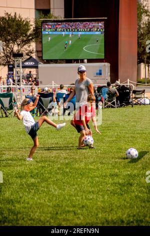 Costa Mesa, Kalifornien, USA. 15. Juli 2018. Während das Publikum tagsüber ein Fußballspiel der Weltmeisterschaft auf einem riesigen Fernsehbildschirm im Freien in Costa Mesa, CA, ansieht, spielt eine Mutter das Spiel mit ihren Kindern auf einem nahe gelegenen Spielfeld. (Kreditbild: © Spencer Grant/ZUMA Press Wire) NUR REDAKTIONELLE VERWENDUNG! Nicht für den kommerziellen GEBRAUCH! Stockfoto