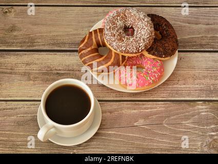 Eine Tasse heißer schwarzer Kaffee und ein Teller mit einem Haufen frischer Donuts in mehrfarbiger Glasur auf einem Holztisch. Draufsicht, flach liegend. Stockfoto