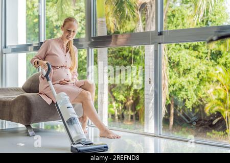 Junge schwangere Frau putzt gern ihr Haus. Einfache Reinigung mit einem kabellosen Staubsauger. Schwangere Frau reinigt Boden mit Handstaubsauger Stockfoto