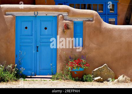 Rancho de Taos, New Mexico, USA - 24. Juli 2023: Eine blau gestrichene Tür und Rollläden im Kontrast zu den braunen lehmwänden eines Privathauses. Stockfoto