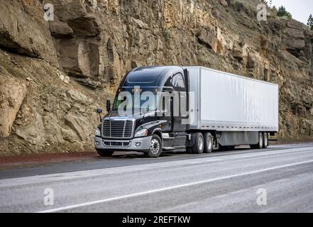 Industrieller schwarzer Sattelschlepper mit hoher Kabine für Lkw-Fahrer, der gewerbliche Fracht in einem trockenen Auflieger transportiert, der auf der Straße steht Stockfoto