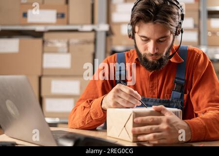 Männlicher Lagerarbeiter schreibt eine Adresse auf eine Kiste, während er vor einem Laptop mit einer Datenbank sitzt Stockfoto