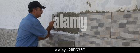 Bild eines Handwerkers, der mit einem Pfannenwender den Klebstoff ausstreut, um die Fliesen an einer Hauswand zu verlegen. Heimarbeit und Hausrenovierung. Banner Stockfoto