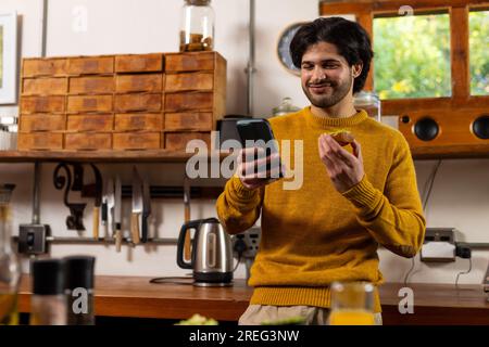 Indischer Mann, der Sandwich mit Avocado isst und zu Hause in der Küche ein Smartphone benutzt Stockfoto
