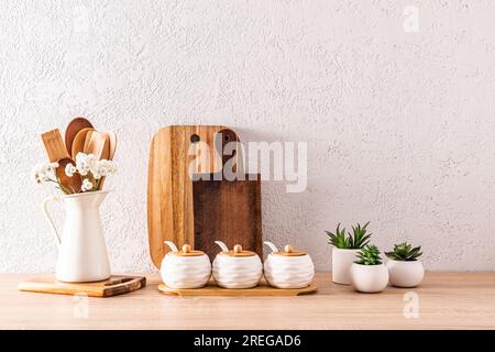 Schneiden von Holzbrettern, Gewürzgläsern, Holzlöffeln in einer Kanne auf einer hölzernen Arbeitsfläche mit Topfblumen. Gemütliches Haus Stockfoto