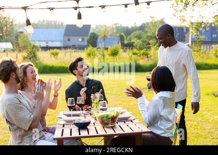 Eine überschwängliche Szene auf einer Gartenparty, bei der ein Schwarzer seiner multiethnischen Gruppe von Freunden aufregende Neuigkeiten überbringt. Die Atmosphäre ist voller Freude und Aufregung, während seine Freunde jubeln und in die Hände klatschen und ihm herzlich zu seinem wohlverdienten Erfolg gratulieren. Fröhliche Feier auf einer Garden Dinner Party, während ein Schwarzer gute Nachrichten teilt. Hochwertiges Foto Stockfoto