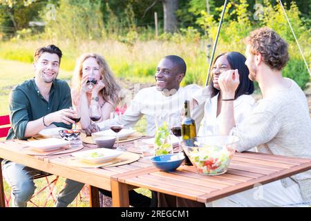 Eine lebendige und vielfältige abendliche Gartenparty, bei der sich eine Gruppe von Millennials am Esstisch versammelt, lebhafte Gespräche führt und die Gesellschaft des anderen genießt. Lachen und Freude füllen die Luft, während sie sich verbinden, und schaffen unvergessliche Erinnerungen in dieser herrlichen Umgebung. Lebhafte abendliche Gartenparty mit multiethnischen Millennials-Freunden, die sich unterhalten und Spaß haben. Hochwertiges Foto Stockfoto