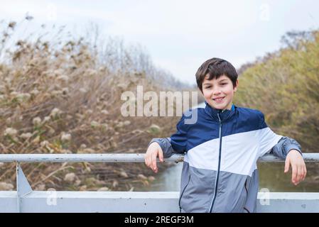 Ein langes Porträt eines jungen Teenagers, der am Geländer auf der Brücke steht, während er in die Kamera schaut und am Fluss lächelt Stockfoto