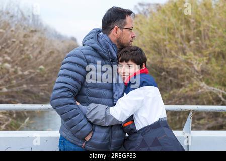 Seitlicher Blick auf einen Vater und Sohn, die sich umarmen, während sie sich über eine Flussbrücke an einem Geländerzaun lehnen Stockfoto