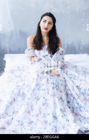Ein Mädchen im Bett in einem weißen Kleid sieht auf grauem Hintergrund weg Stockfoto